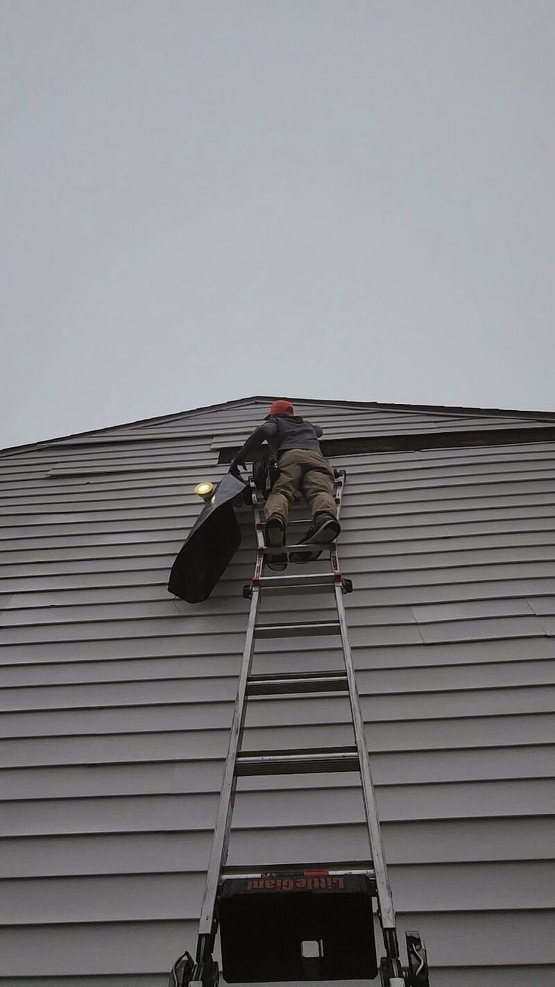 Crew member climbing a ladder to the roofline of a house with grey horizontal siding