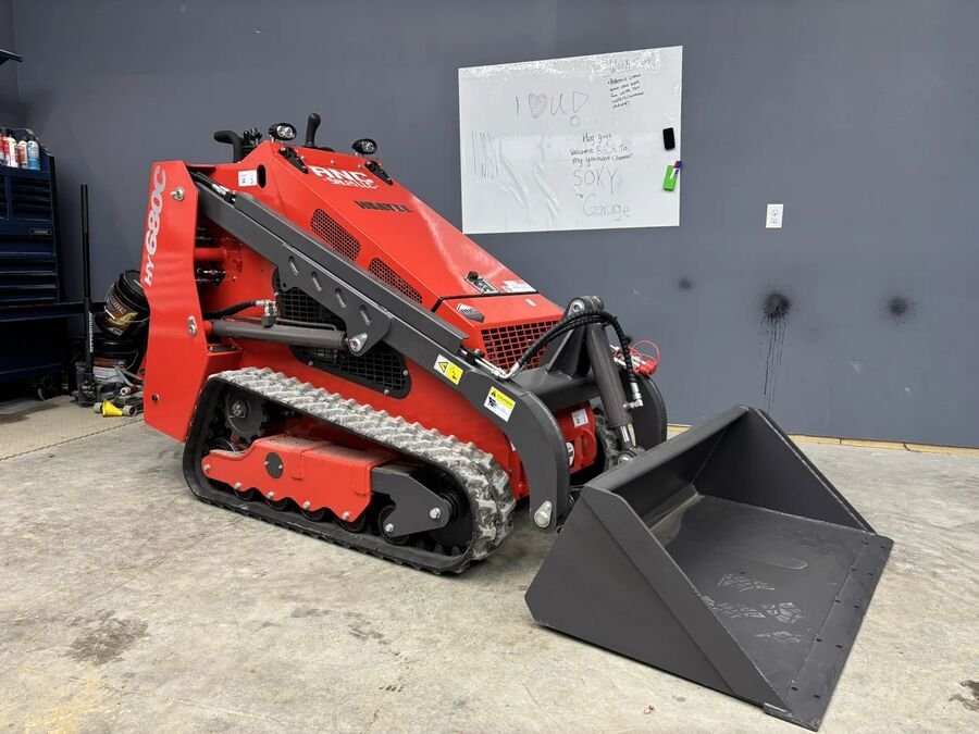 Red Huayee HY680C mini skid steer on tracks with a bucket, in front of a gray shop wall