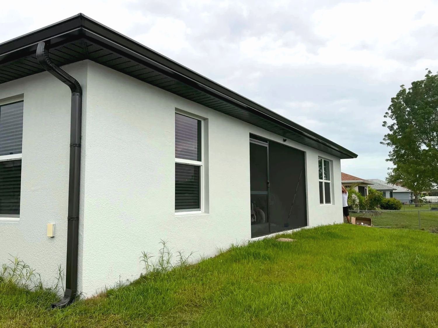 Black seamless gutter and downspout on the corner of a white stucco house, screened lanai at the back, green lawn