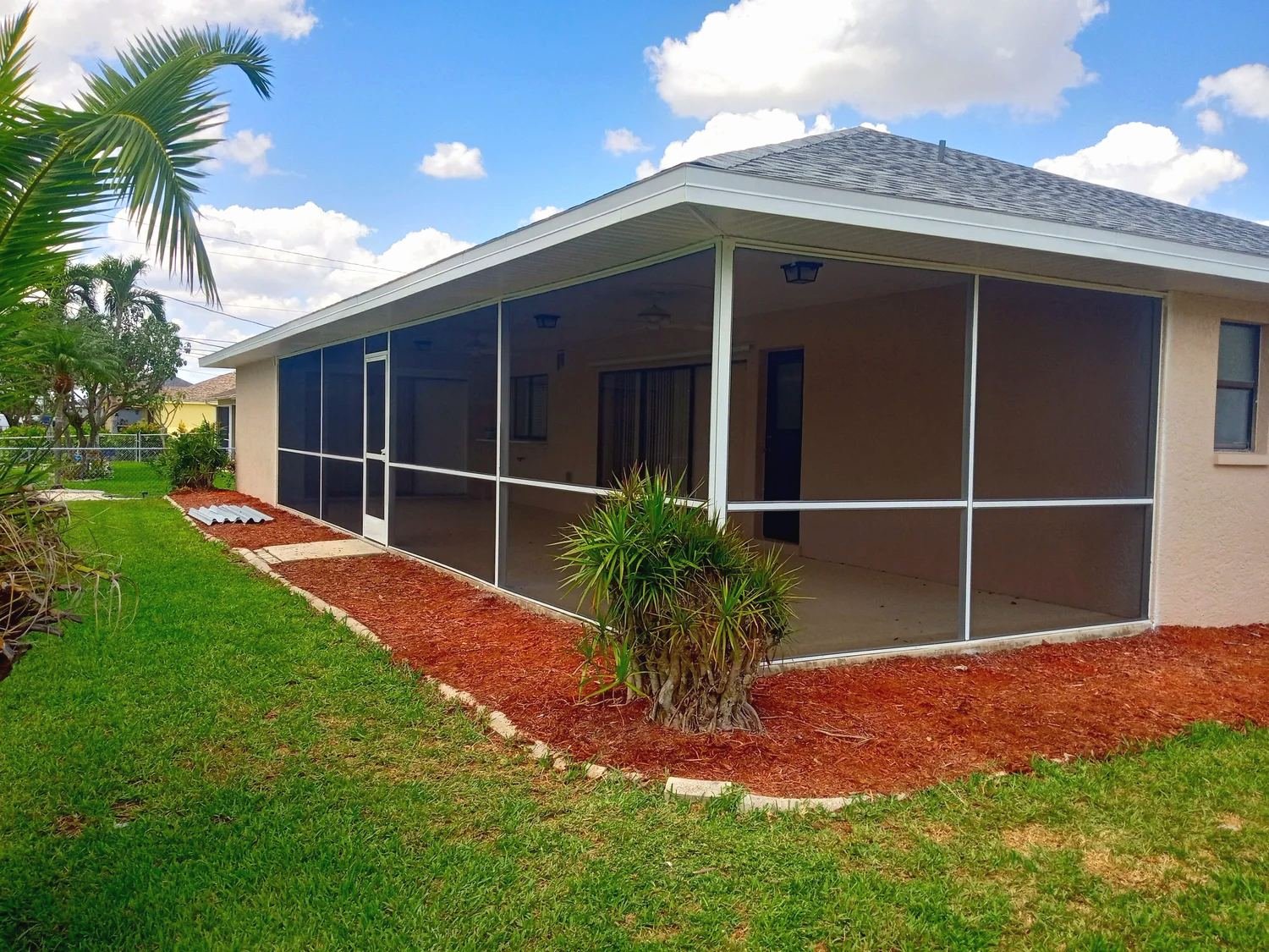 White-framed screen room with a solid insulated roof on the back of a tan house, palm and red mulch bed in front