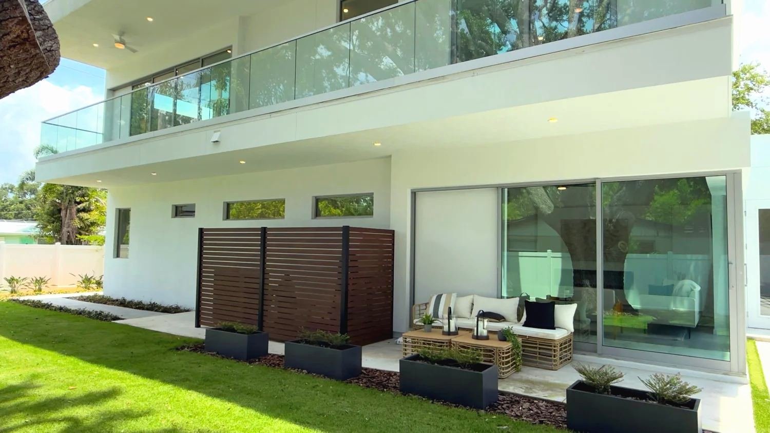 Frameless glass balcony railing on a white modern house above a covered patio with outdoor seating and a wood slat privacy screen