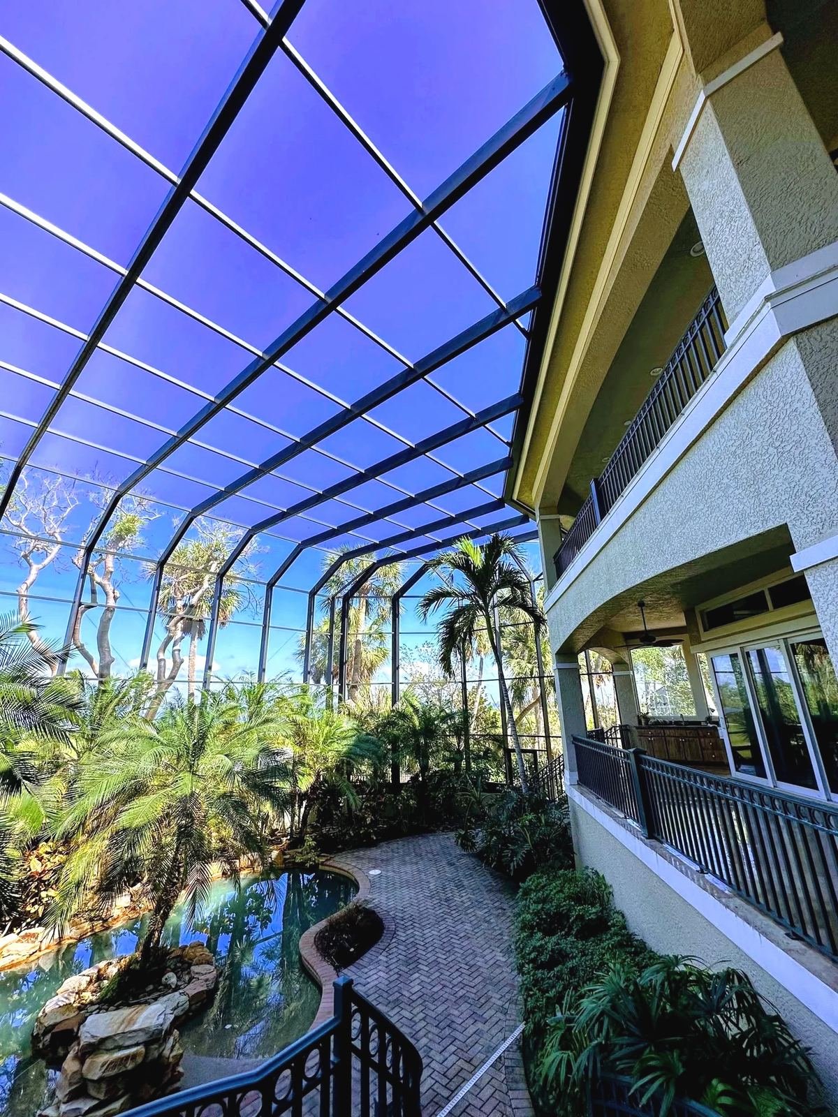 Two-story black pool cage from inside, looking up at the mansard roof panels beside a stucco house with iron balcony railings