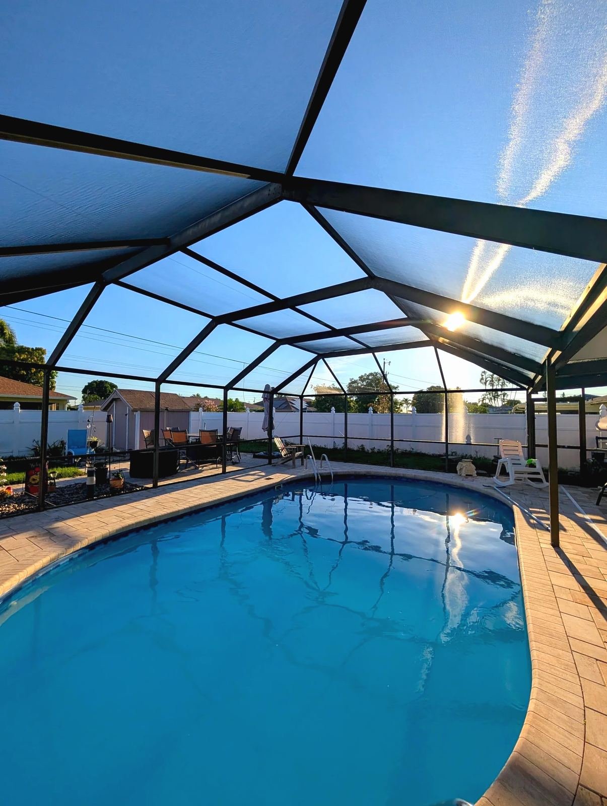 Inside a dark bronze pool cage at sunset, oval pool and paver deck below, white vinyl fence and patio chairs beyond the screen