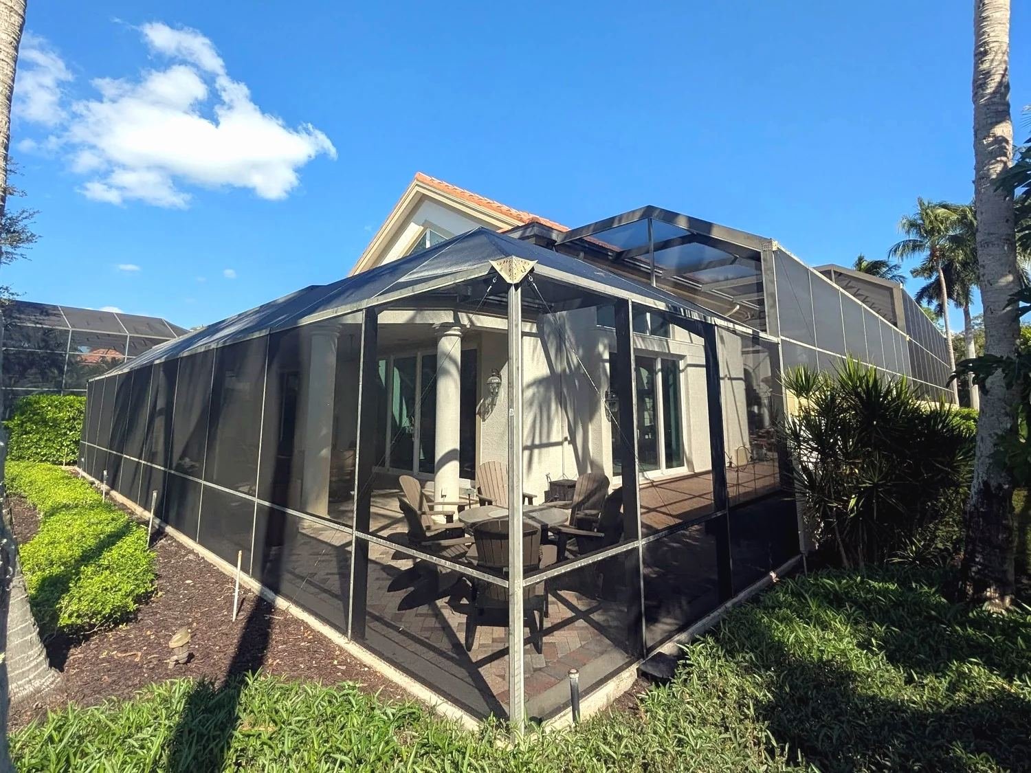 Bronze pool cage with stepped roof heights against a two-story tile-roof house, patio chairs inside, Southwest Florida