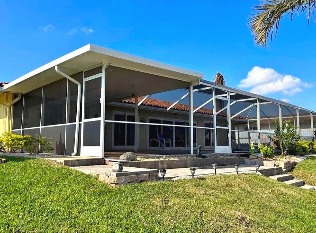 White insulated roof over a lanai joined to a white pool cage, tile-roof house behind, terraced lawn in front